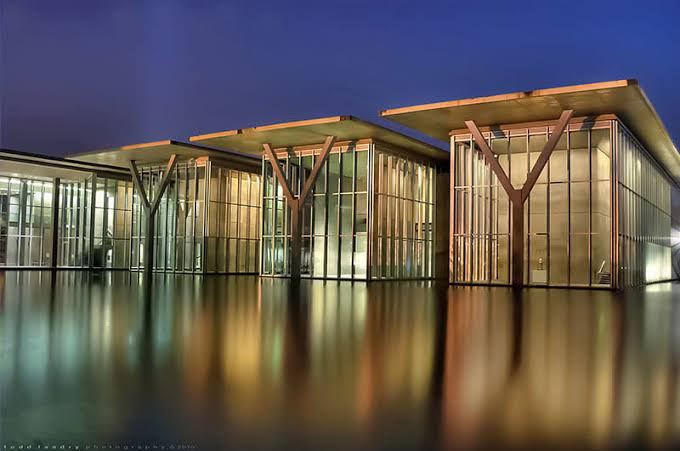 Modern glass buildings with V-shaped supports reflecting on water at night.
